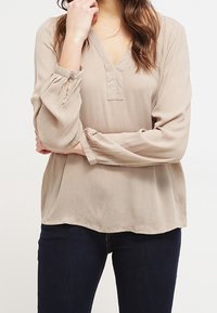 Blouse beige légère avec un col en V, manches longues avec poignets froncés et une coupe décontractée. Fabriquée en tissu doux et texturé.
