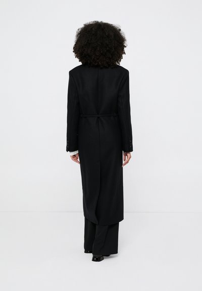 Mulher com cabelo encaracolado, vestindo um casaco longo preto, calças pretas e sapatos pretos, em pé de frente para um fundo branco liso.