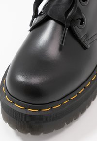 Stivale alla caviglia in pelle nera con finitura lucida, punta rotonda, suola spessa nera e cuciture gialle lungo il bordo. Design con lacci in tessuto.