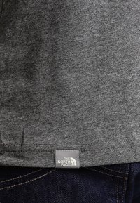Camiseta de algodón gris con una textura suave, que presenta un pequeño parche de logo etiquetado como "The North Face" en el dobladillo, combinada con jeans oscuros.