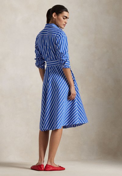 Naine sinise ja valge triibulise pika varrukaga kleidis, millel on kroogitud varrukad, seisab paljajalu neutraalsel taustal ja vaatab üle õla.