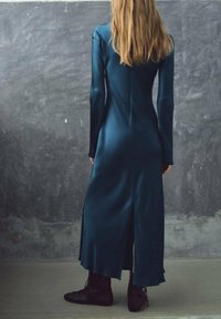 Mouwloze, teal satijnen jurk met een split aan de achterkant, een aansluitende silhouet en naadloos ontwerp. Gecombineerd met zwart gelakte enkellaarsjes.