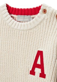 Pull crème tricoté avec un col côtelé rouge, présentant un grand "A" rouge brodé sur le devant et des accents de boutons en bois.
