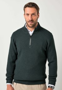 Hombre con barba corta que lleva un suéter de cremallera de canalé en verde oscuro sobre una camisa blanca y pantalones beige, de pie contra un fondo claro y liso.