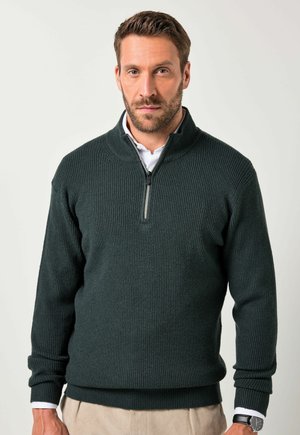 Hombre con barba corta que lleva un suéter de cremallera de canalé en verde oscuro sobre una camisa blanca y pantalones beige, de pie contra un fondo claro y liso.