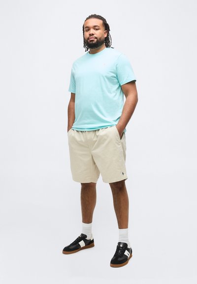 Νεαρός με dreadlocks στέκεται με τα χέρια στις τσέπες, φορώντας γαλάζιο κοντομάνικο μπλουζάκι, μπεζ σορτς, λευκές κάλτσες και μαύρα αθλητικά παπούτσια.