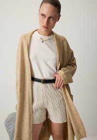 Beżowy kardigan, biały top, kremowe szorty w warkocz, czarny pasek z kwadratową klamrą. Gładki materiał, luźny krój, minimalne detale.