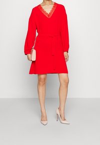 Rotes Kleid mit langen Ärmeln, V-Ausschnitt mit Spitzenbesatz, tailliert mit einem passenden Gürtel. Kombiniert mit hellrosa High Heels.