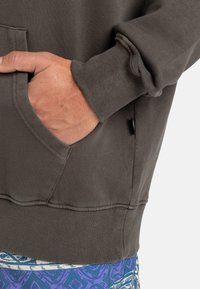 Grå hettegenser med teksturert stoff, med frontlomme og ribbestrikkede mansjetter. Synlig sømdetalj og en sideetikett som aksent.