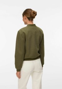 Femme avec les cheveux en chignon, portant un pull à manches longues vert olive et un jean blanc, debout de dos face au spectateur sur un fond blanc.