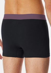 Boxer in tessuto nero con elastico viola in vita. Presenta un design aderente e una piccola apertura nella parte anteriore. Orli cuciti curati.