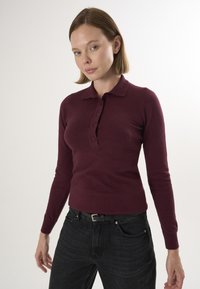 Langærmet polo i bordeaux med knaplukning, lavet af blødt stof, kombineret med mørke denimjeans og et sort bælte.