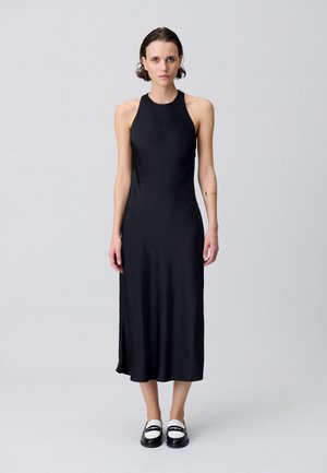 Robe midi sans manches noire en tissu lisse et mat ; présente une encolure haute, une fente sur le côté et une silhouette ajustée. Assortie avec des chaussures noires et blanches.