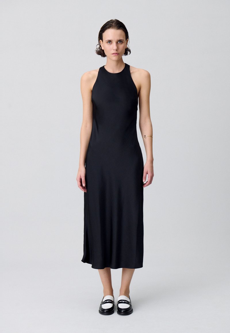 Robe midi sans manches noire en tissu lisse et mat ; présente une encolure haute, une fente sur le côté et une silhouette ajustée. Assortie avec des chaussures noires et blanches.