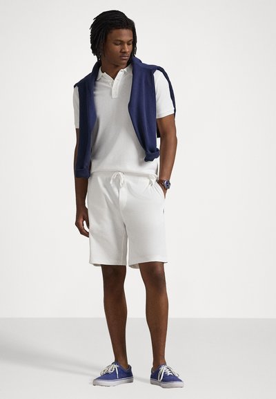 Hvite shorts, hvit polo-skjorte og en marineblå genser drapert over skuldrene. Blå joggesko fullfører antrekket. Uformell og komfortabel passform.