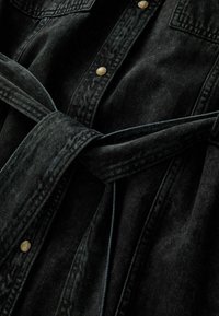 Zwarte spijkerbloes met een knoop in de taille, voorzien van twee borstzakken en goudkleurige knopen, en met een licht gestructureerde stof.