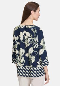 Kwiatowa bluzka w kolorze granatowym z dużym zielonym i kremowym wzorem kwiatowym. Posiada rękawy o długości trzech czwartej oraz falbaniasty brzeg. Gładka faktura materiału.