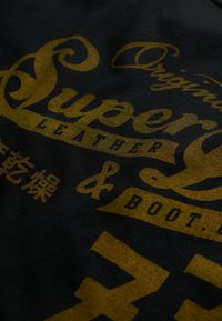 Sort stof med fed, gylden tekst i dekorativ skrift og symboler. Designet inkluderer ”Super Leather & Boot Co.” accenter.