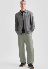 Zwart-wit geblokte overhemd over een zwart T-shirt, gecombineerd met losse groene broeken en zwarte sneakers met een gumkleurige zool.