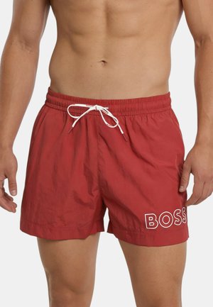 Torace maschile e parte superiore delle gambe con pantaloncini da bagno rossi con coulisse bianca e logo "BOSS" sulla gamba sinistra.