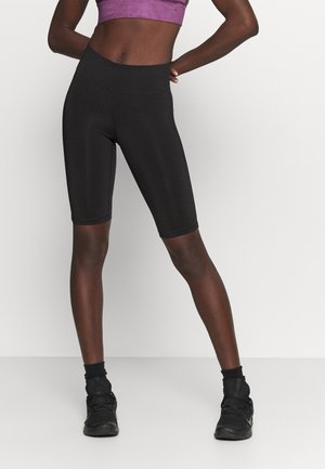 Person trägt schwarze knielange Sportshorts, schwarze Sneakers und ein lila Sportoberteil, steht mit den Händen in der Hüfte.