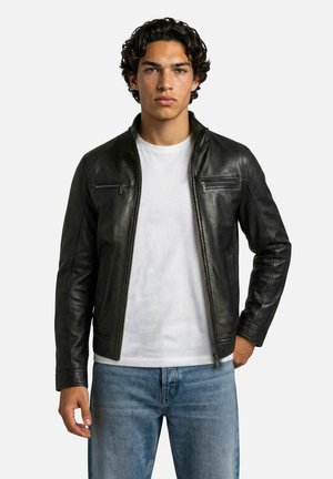 Jonge man met krullend donker haar, die een zwarte leren jas draagt over een wit T-shirt en blauwe spijkerbroek, staand tegen een effen achtergrond.
