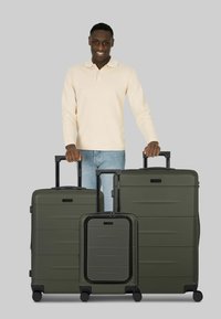 Quattro valigie rigide verdi di varie dimensioni con superfici lisce e basi nere con ruote, posizionate davanti a una persona con un maglione chiaro.