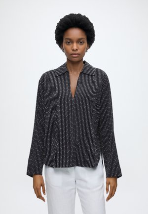 Femme aux cheveux courts et bouclés portant une chemise noire à manches longues avec des rayures verticales blanches et un pantalon blanc, debout devant un fond uni.