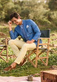 Blazer a quadri blu chiaro sopra un polo blu a maglia, abbinato a pantaloni gialli e scarpe slip-on marroni. Ambientazione all'aperto con erba verde.