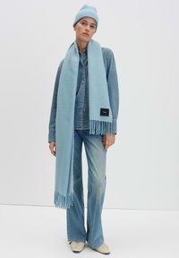 Lichtblauwe gebreide beanie en sjaal, gecombineerd met een spijkerblouse, uitlopende jeans en crèmekleurige schoenen. De sjaal heeft franjes en een zwart label.