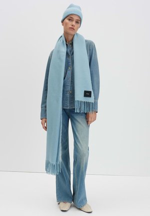 Helle blaue Strickmütze und Schal, kombiniert mit einem Jeanshemd, weit geschnittenen Jeans und cremefarbenen Schuhen. Der Schal hat einen Fransensaum und ein schwarzes Etikett.