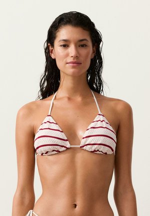 Femeie cu păr închis la culoare ud, purtând un top de bikini alb cu dungi roșii orizontale pe un fundal simplu.