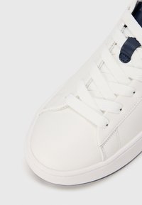 Biela teniska s hladkým koženým povrchom, vybavená bielymi šnúrkami, námorníckym modrým akcentom blízko vrchu a zaoblenou gumovou podrážkou.