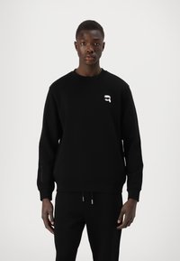 Črna majica s dolgimi rokavi, rebrastimi manšinami in okroglim izrezom. Ima majhen bel logo na prsih in je izdelana iz mehkega blaga.