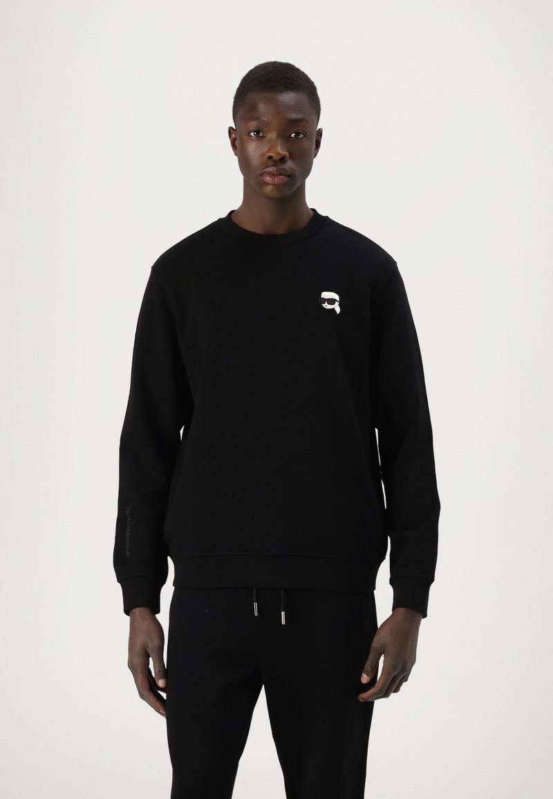 Črna majica s dolgimi rokavi, rebrastimi manšinami in okroglim izrezom. Ima majhen bel logo na prsih in je izdelana iz mehkega blaga.