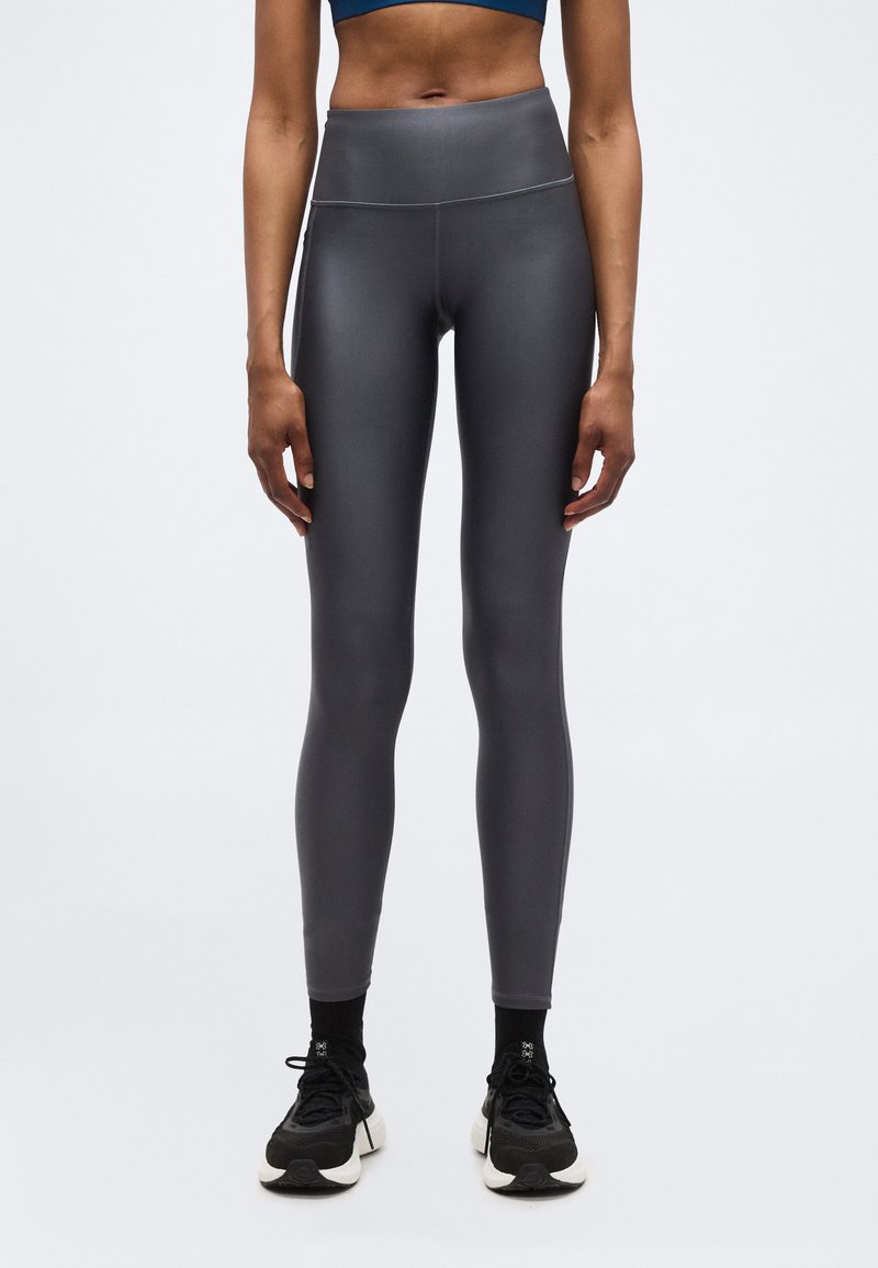 Γυναίκα που φοράει ψηλόμεσα σκουρόχρωμα γκρι κολάν, μαύρες κάλτσες και μαύρα αθλητικά παπούτσια, στέκεται μπροστά σε ένα απλό λευκό φόντο.