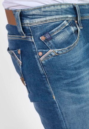 Jeans de denim en un tono azul medio con acentos desgastados, que presentan un diseño clásico de cinco bolsillos, remaches de cobre y una etiqueta de cuero marrón.