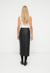 Svart lær midi skjørt med en strømlinjeformet design, med en baksliss og jevn tekstur. Kombinert med en hvit ermeløs topp og mørke sko.