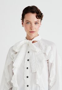 Blusa blanca con detalle de encaje con volantes, cuello con lazo, botones de flor negra en contraste, tela texturizada y ribete de encaje intrincado a lo largo de las mangas.