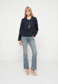 Jūras flotei piederīga jaka ar teksturētu audumu, īsa, apgriezta piegriezuma un pogām; saskaņota ar gaiši ziliem izvērstiem džinsiem un bēšām augstpapēžu kurpēm. Zem tās valkāts balts tops.