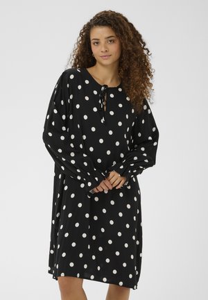 Kvinde med krøllet hår iført en sort knælængde kjole med hvide prikker og lange, puffede ærmer, stående med hænderne foldet.