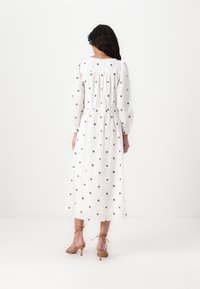 Vit långärmad klänning med svart blommönster, samlad midja och flödande kjol. Kombinerad med tan färgade klackar för en fullständig look.