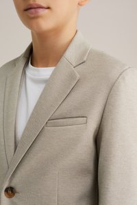 Bežinis švarkas su tekstūruota medžiaga, viena ruda saga ir krūtinės kišenė. Dėvimas ant balto marškinėlių su apvalia kaklo iškirpte.