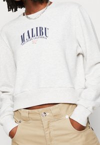 Světlá šedá zkrácená mikina s potiskem "MALIBU SOUTHERN CALIFORNIA" v navy modré. Má žebrovaný výstřih a manžety. Doplňuje ji béžové kalhoty.