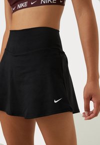 Femme portant une jupe de sport noire Nike et un soutien-gorge de sport bordeaux Nike, debout avec le bras détendu le long du corps sur un fond uni.