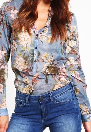 Vrouw draagt een blauwe bloemenprint blouse met knopen, gecombineerd met een hooggetailleerde blauwe spijkerbroek, waarbij de torso en bovenbenen zichtbaar zijn.
