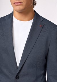 Granatowy żakiet o fakturowym wykończeniu, dopasowany, zapinany na jeden guzik. Noszony na białej koszulce z okrągłym dekoltem. Posiada małe czerwone logo na klapie.