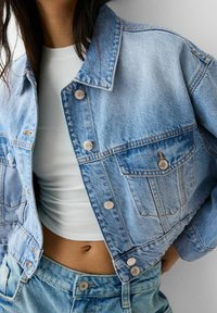 Hellblaue Jeansjacke im Cropped-Design mit silbernen Knöpfen, Brusttasche und ausgefransten Kanten, kombiniert mit einem weißen, enganliegenden Top.