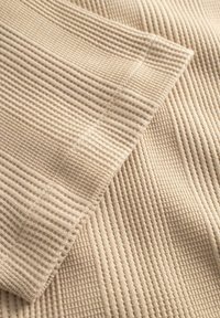 Beige ribbestrikket stoff brettet med synlig mansjettssøm og mykt teksturert mønster på en nøytral bakgrunn.