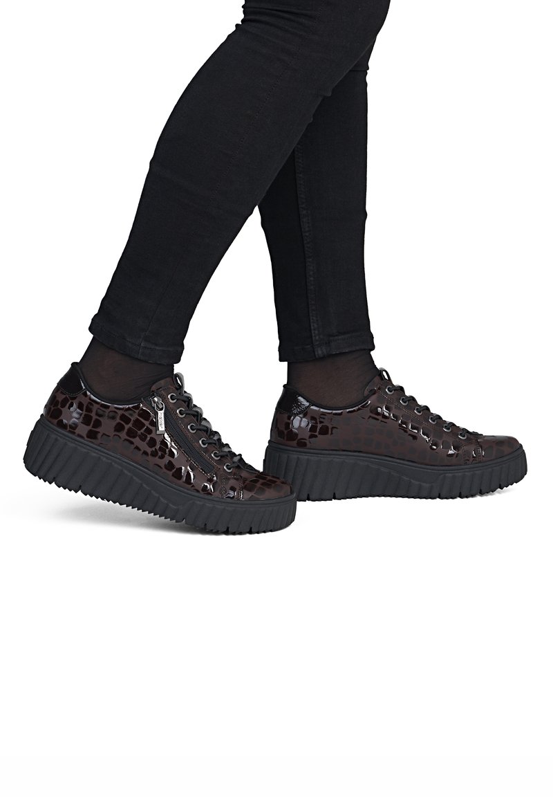 Personne portant des baskets à plateforme brillantes marron foncé à motif crocodile avec des semelles noires et un jean skinny noir.
